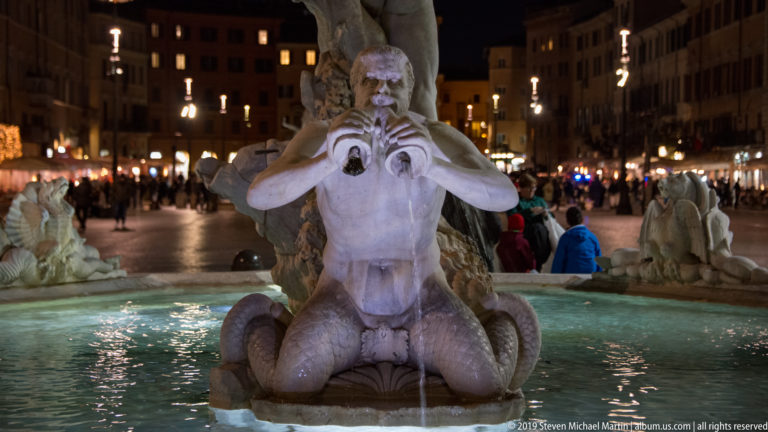 Front view of a kneeling male fountain figure holding an object at his chest, with Piazza Navona crowded behind.