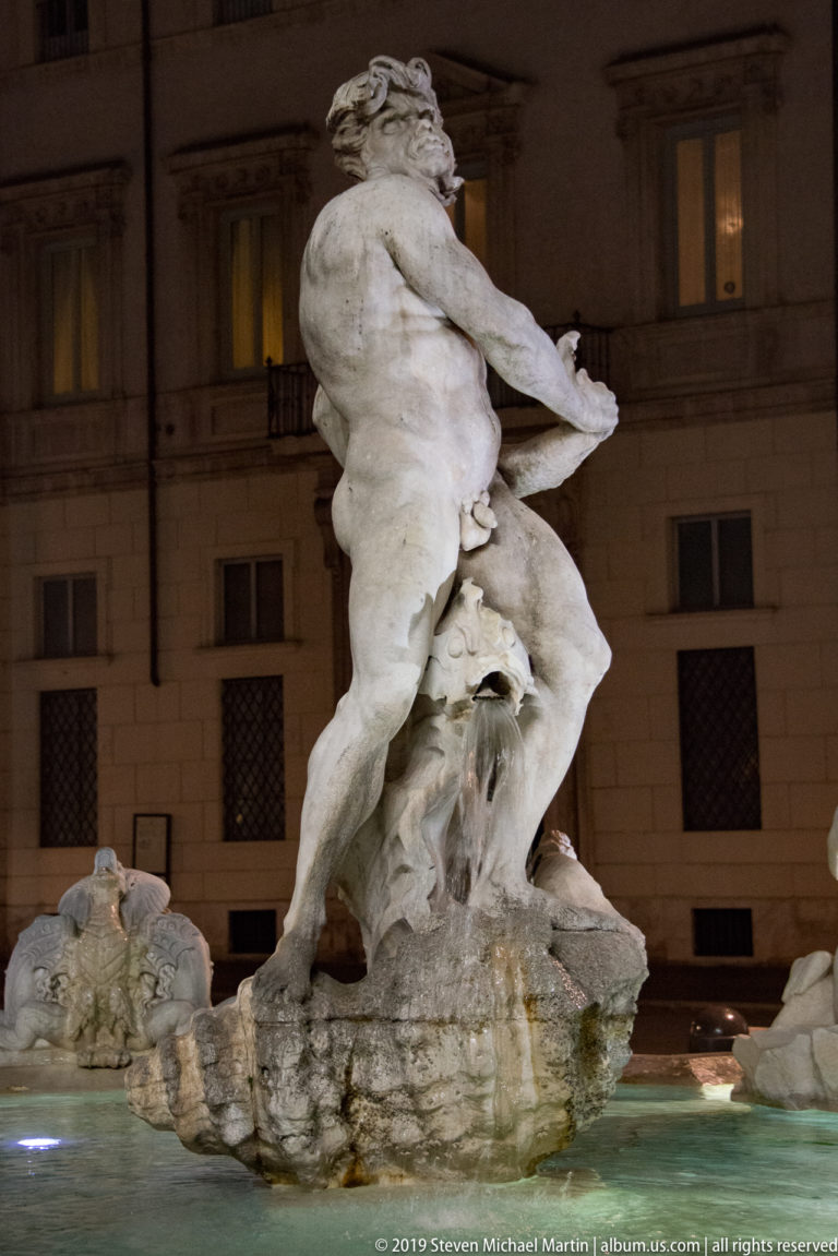 Side view of Neptune’s muscular marble figure wrestling a sea creature above the Fountain of Neptune basin at night.