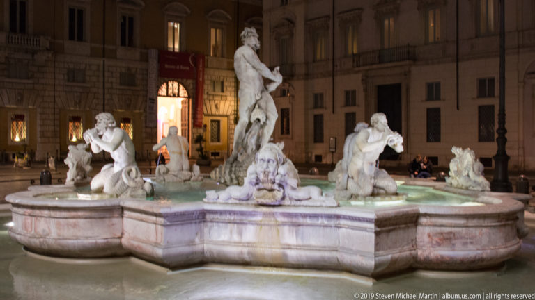 Wide nighttime view of the Fountain of Neptune with multiple marble figures around its basin in Piazza Navona.