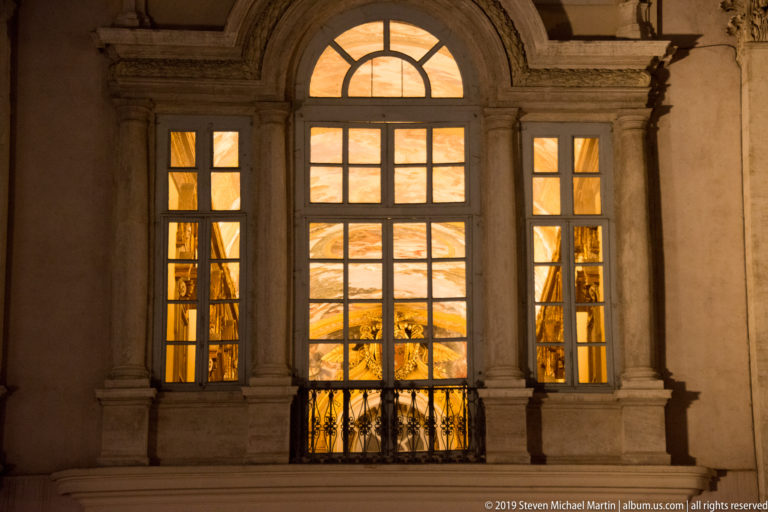 Large arched multi-pane window with a small iron balcony glowing warmly in a pale stone façade at night.