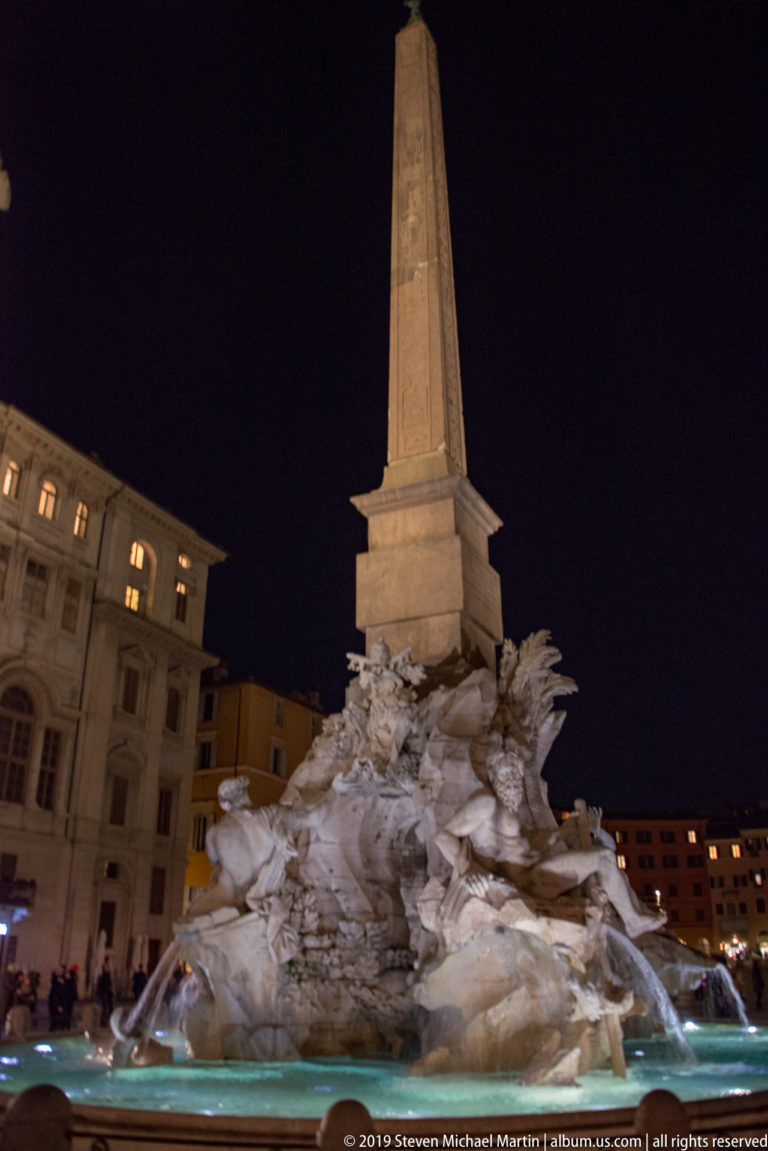 Side view of the Fountain of the Four Rivers with tall obelisk, rocky sculptural base, and illuminated basin at night.