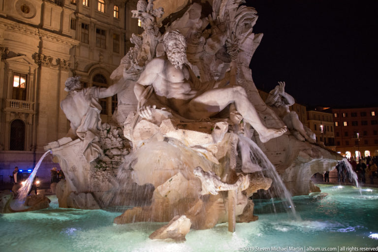 Large reclining river-god figure on rocky fountain masonry with several streams of water falling into an illuminated basin.