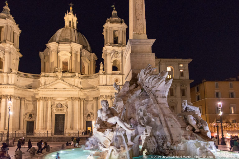 Illuminated Sant’Agnese in Agone behind the rocky Fountain of the Four Rivers and its tall obelisk at night.