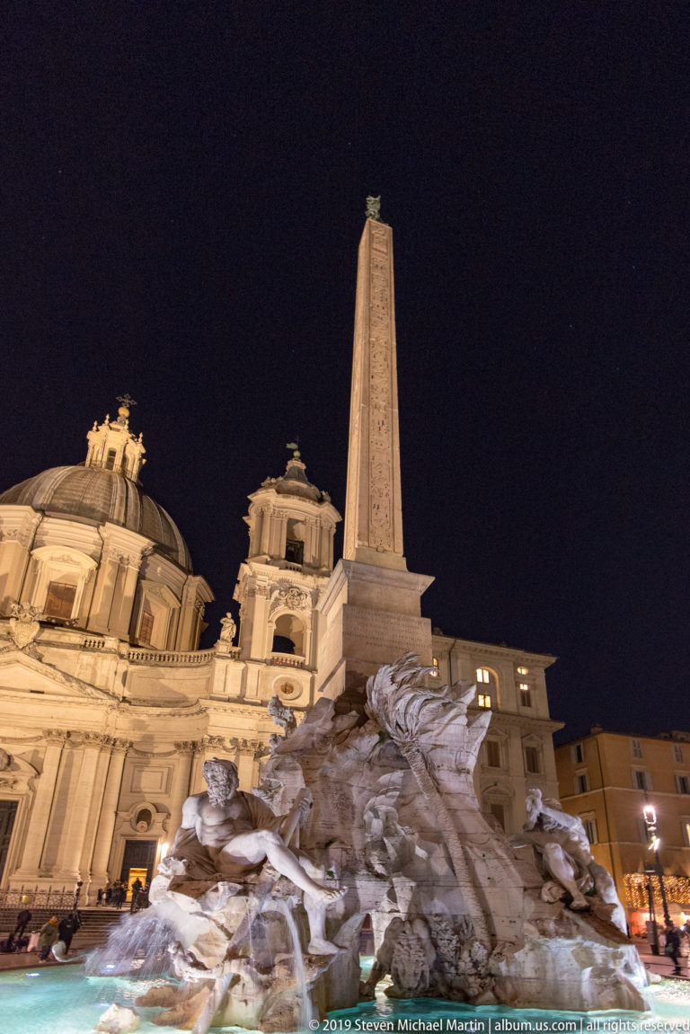 Wide nighttime view of the Fountain of the Four Rivers, obelisk, and illuminated Sant’Agnese in Agone in Piazza Navona.