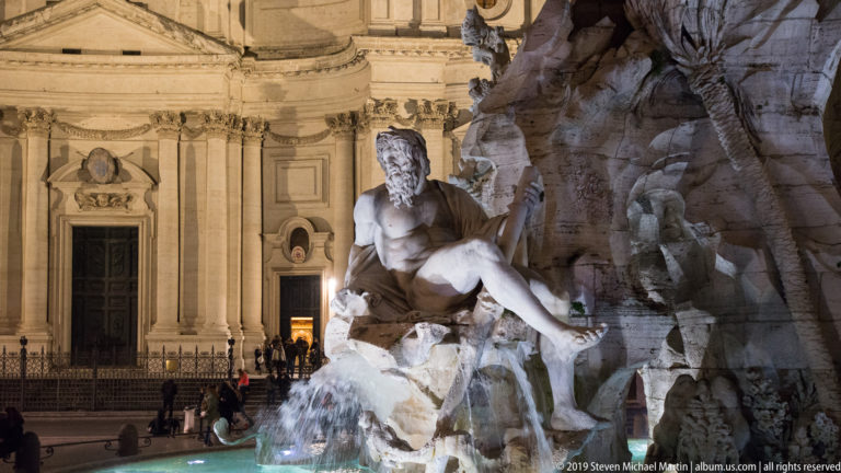 Reclining marble river figure on the Fountain of the Four Rivers with rockwork, flowing water, and Sant’Agnese behind.