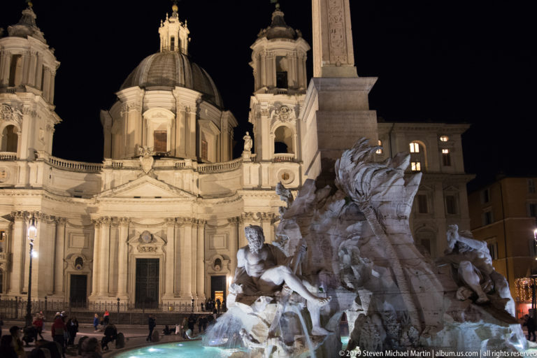 Fontana dei Quattro Fiumi and its obelisk before the illuminated dome and façade of Sant’Agnese in Agone at night.