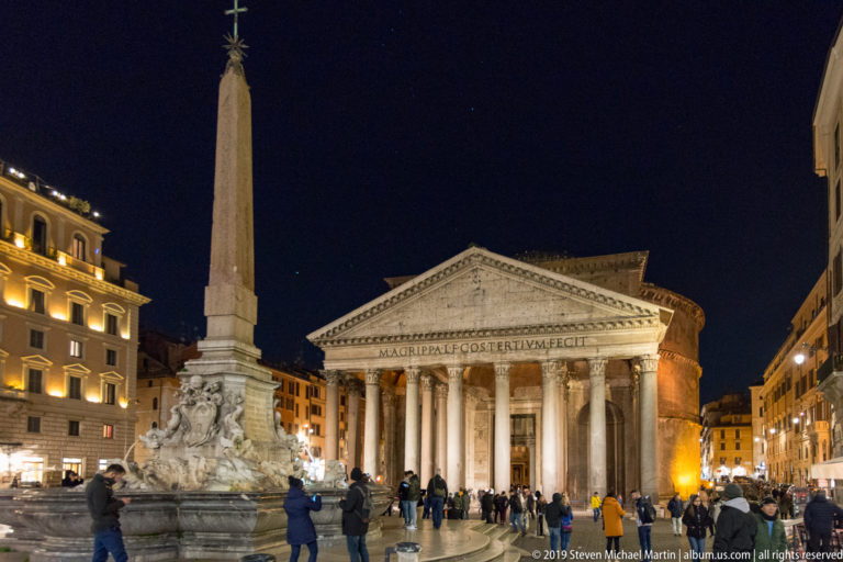 Pantheon and obelisk-topped Fontana del Pantheon illuminated at night with visitors gathered across Piazza della Rotonda.