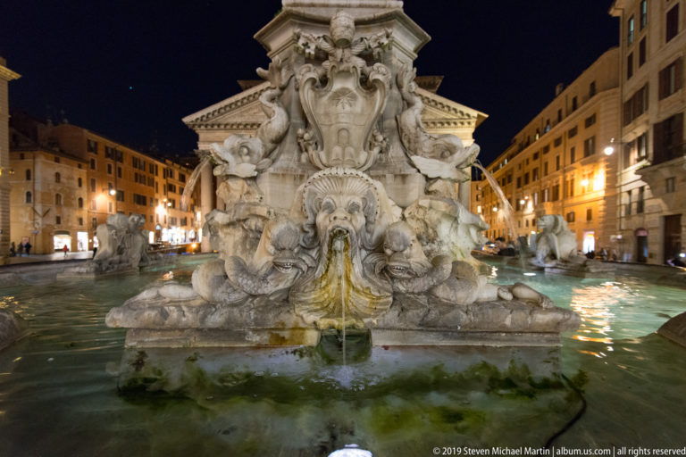 Close frontal view of a carved mask and water spout on the Fontana del Pantheon, with the piazza behind.