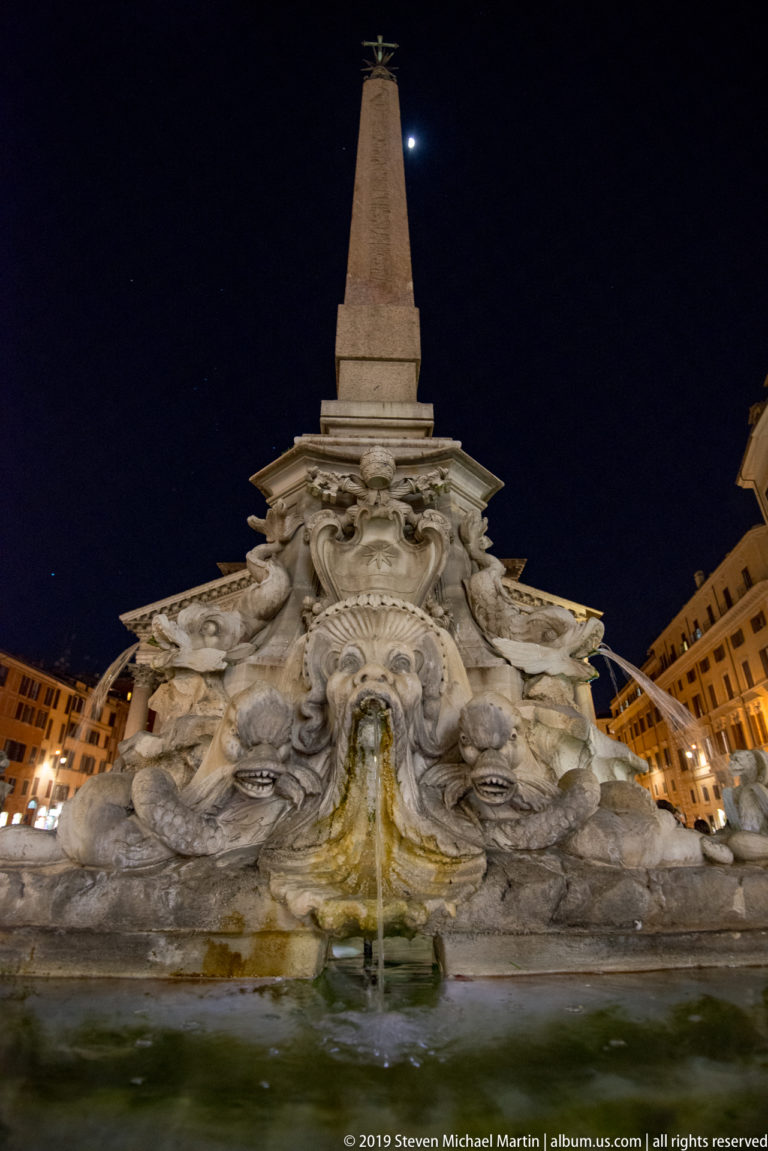 Illuminated Fontana del Pantheon with carved masks and rockwork below the tall obelisk against a dark night sky.