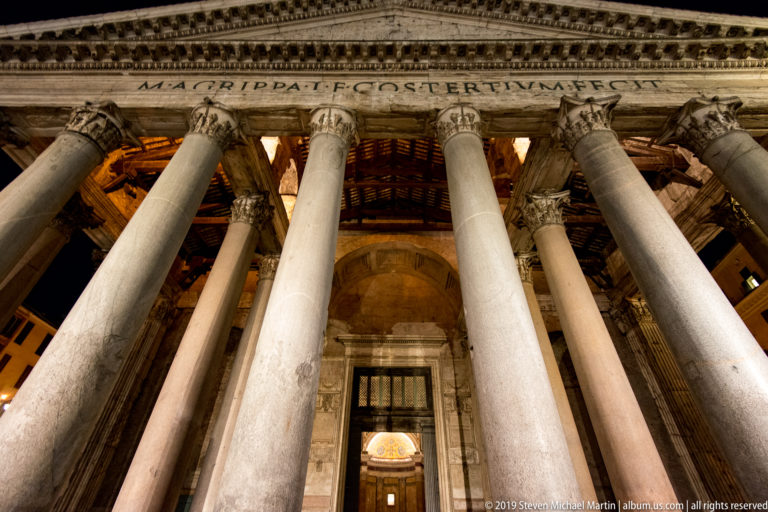 Low-angle nighttime view through the Pantheon’s tall Corinthian columns toward its bronze entrance doors and illuminated portico.