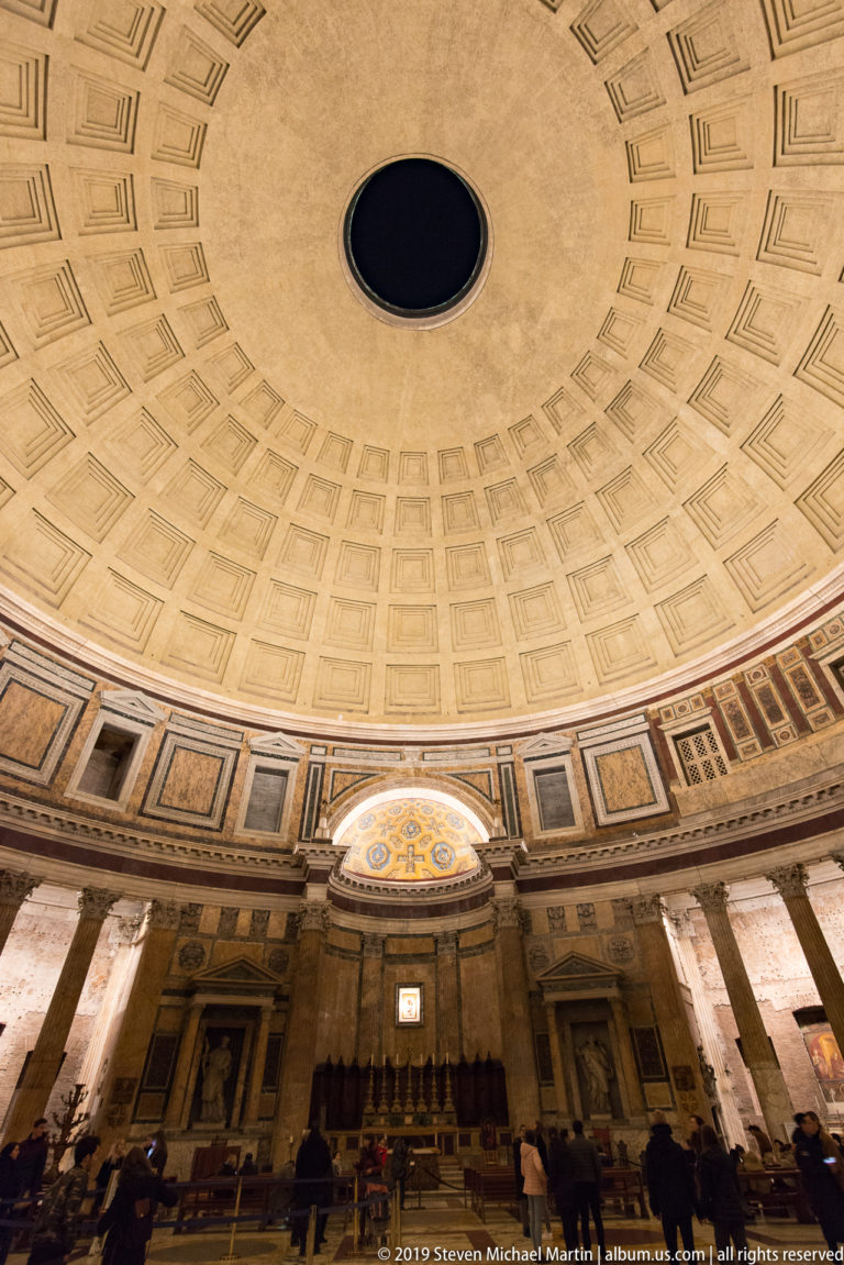 Broad Pantheon interior with coffered dome and oculus above the sanctuary, marble wall articulation, columns, and visitors below.