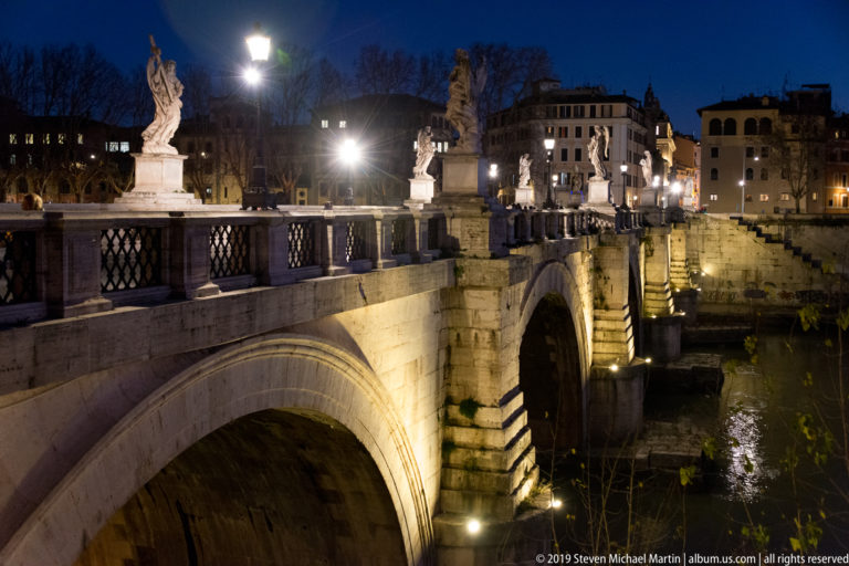 Oblique nighttime view of Ponte Sant’Angelo with illuminated stone arches, marble statues, and reflections in the Tiber.
