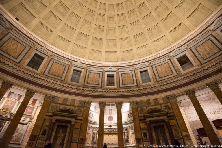 Oblique Pantheon interior with coffered dome above marble wall panels, tall columns, and statue niches around the rotunda.