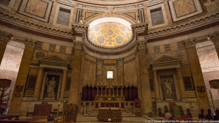 Pantheon sanctuary centered beneath decorated apse, with altar, columns, marble wall panels, and statue-filled niches on both sides.