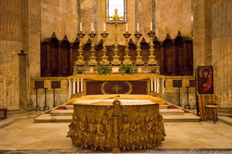 Pantheon sanctuary with sculpted altar frontal, white altar cloth, gilded candlesticks, dark choir stalls, and tall marble columns.