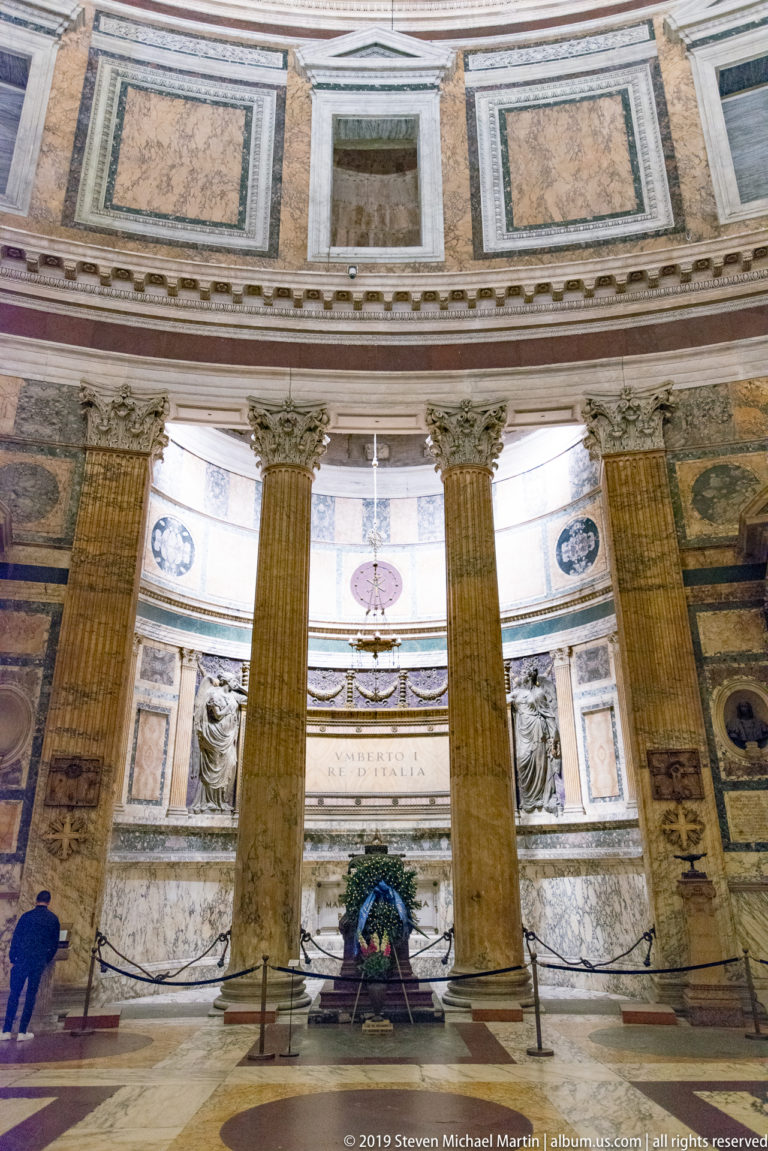 Curved Pantheon interior wall with colored marble panels, columns, niches, cornices, chapel furnishings, and visitors along the perimeter.