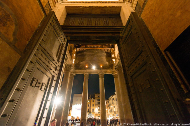 Open paneled bronze doors of the Pantheon framing portico columns, bright piazza lights, and a crowd outside at night.