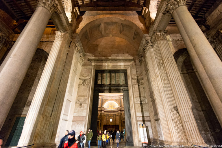 Massive Pantheon columns frame the bronze entrance doors, with visitors gathered beneath the deep illuminated portico.