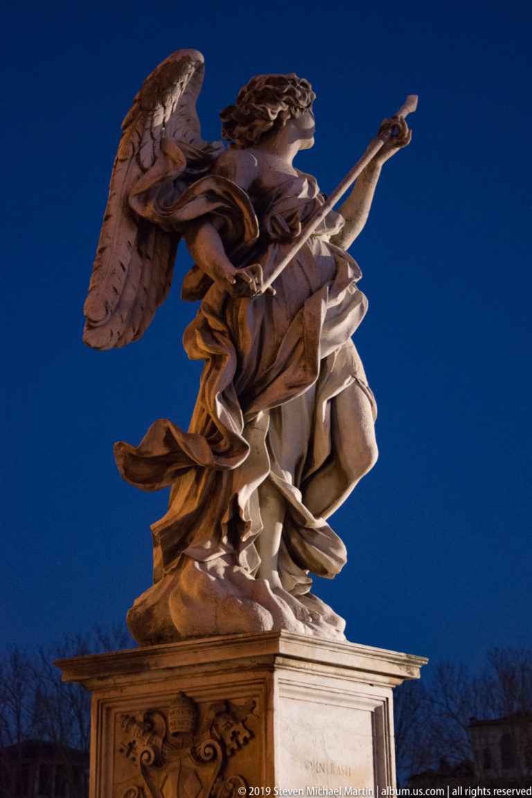 Floodlit marble Angel with the Lance by Domenico Guidi on Ponte Sant’Angelo, holding a long spear across swirling drapery.