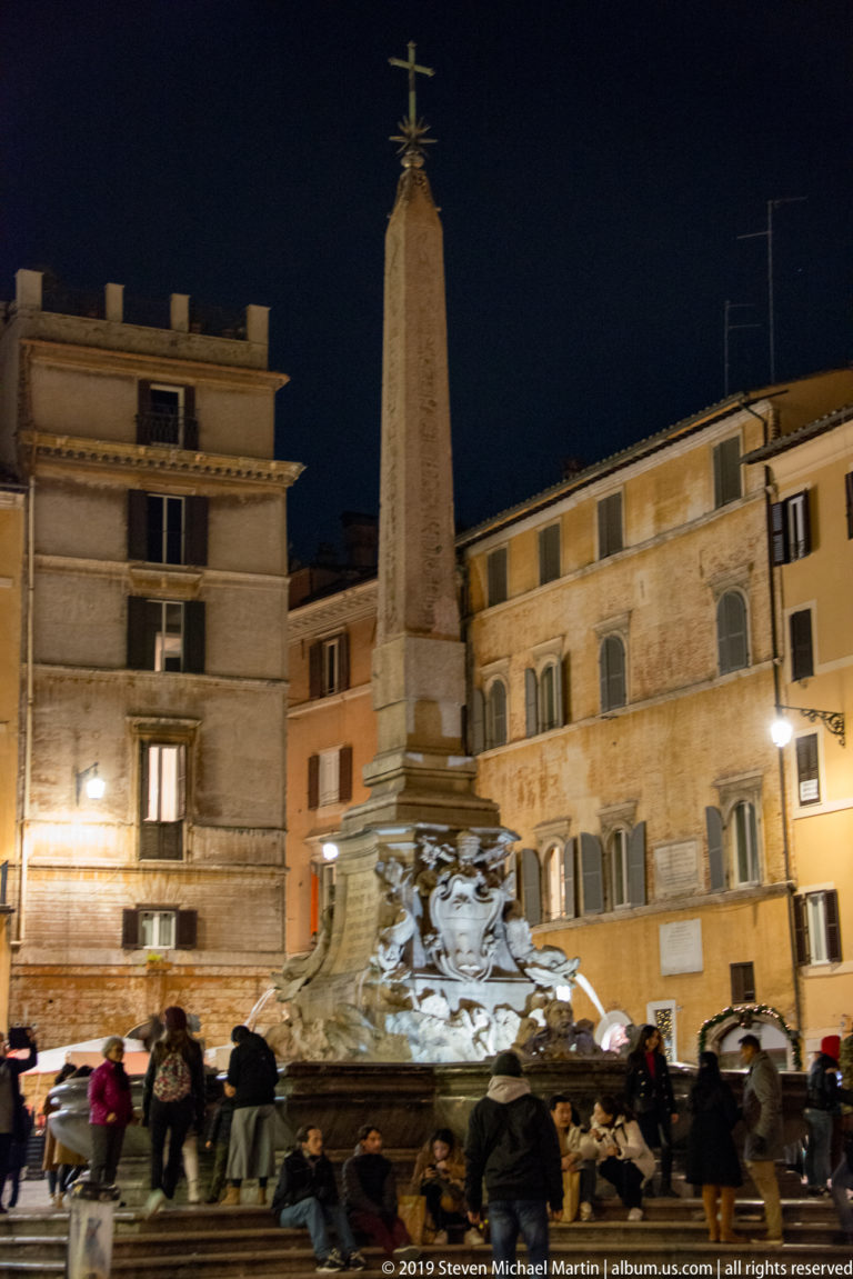 Obelisk rising above the Fountain of the Four Rivers at night, with sculpted fountain base, crowds, and surrounding Piazza Navona façades.