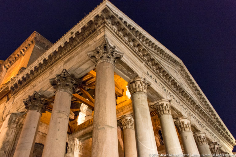 Low-angle night view of the Pantheon portico with massive Corinthian columns, carved capitals, triangular pediment, and timber roof behind.