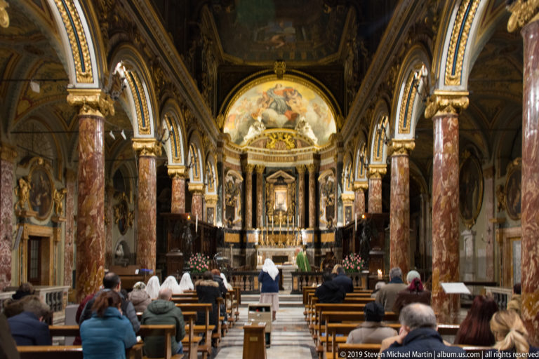Ornate church nave with seated worshippers, marble columns, gilded decoration, painted apse, and clergy gathered near the high altar.