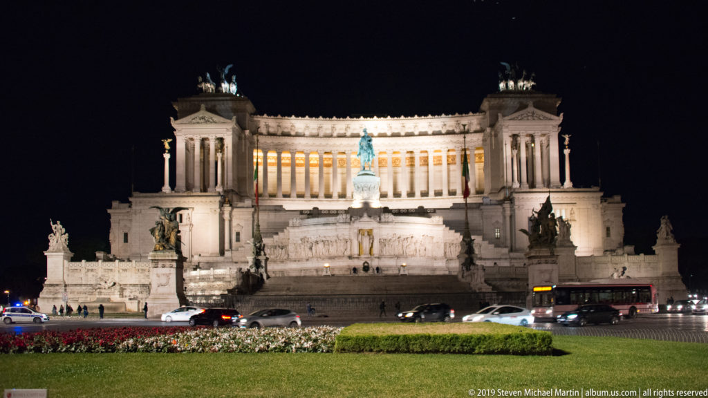 Floodlit Vittoriano at night beyond lawn, flowers, and passing cars, with central statue and rooftop quadrigas clearly visible.