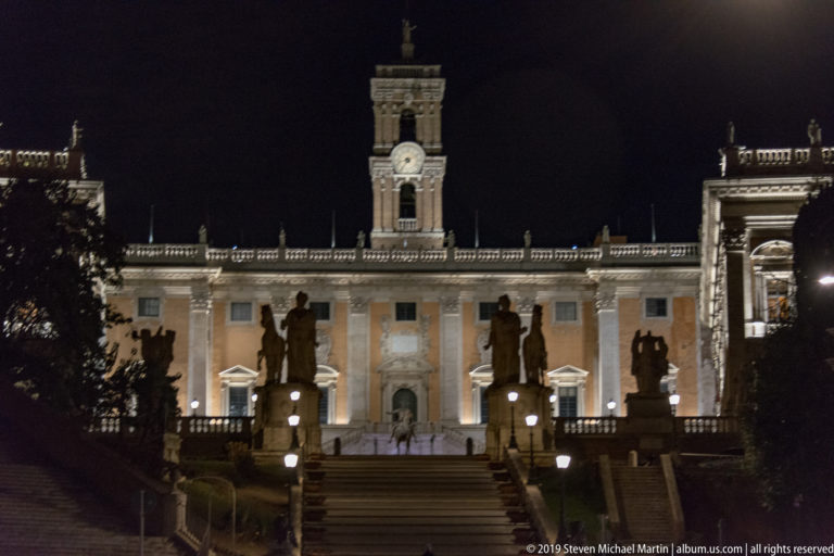 Illuminated Palazzo Senatorio and clock tower above a broad stair approach, with dark statues and balustrades in the foreground.