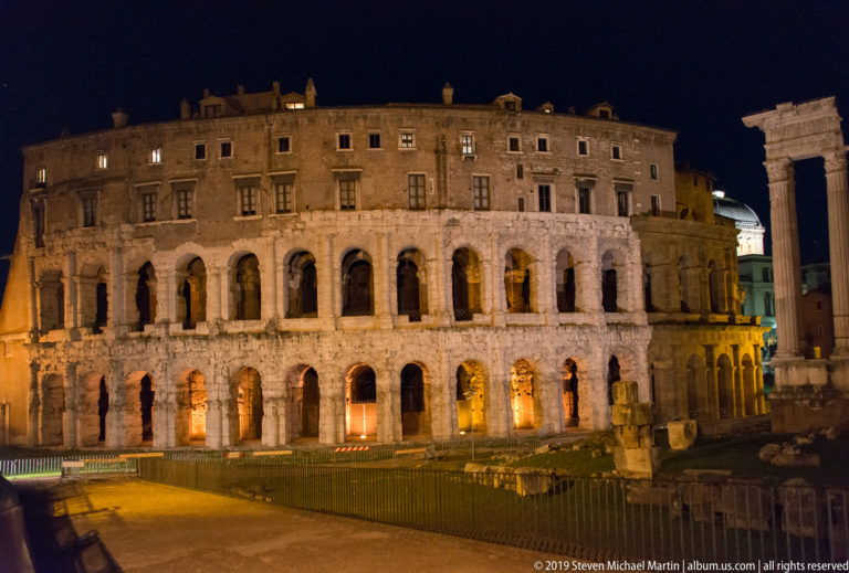 Illuminated curved façade of the Theatre of Marcellus, with ancient arcades below later upper stories and fenced ruins in front.