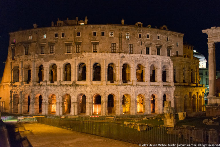 Wide night view of the Theatre of Marcellus with illuminated ancient arcades, later upper stories, and archaeological remains in front.