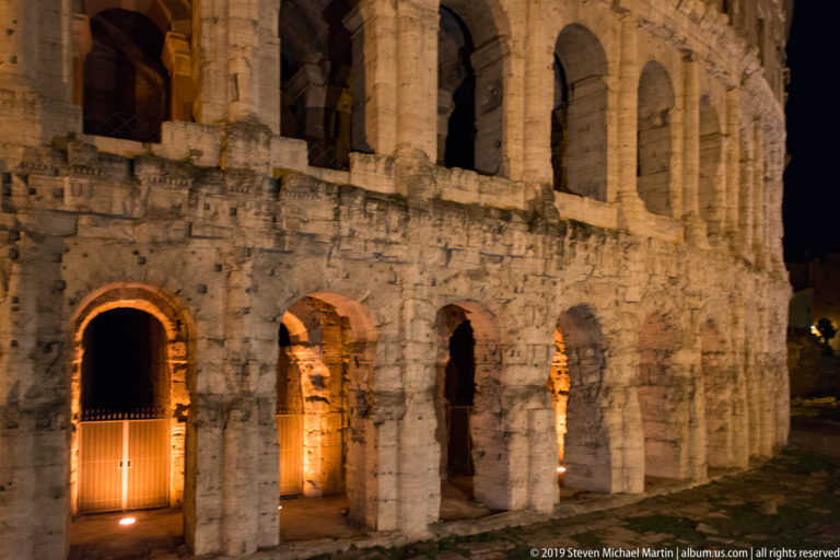 Close night view of the Colosseum’s lower arched openings and weathered masonry, lit warmly from within.