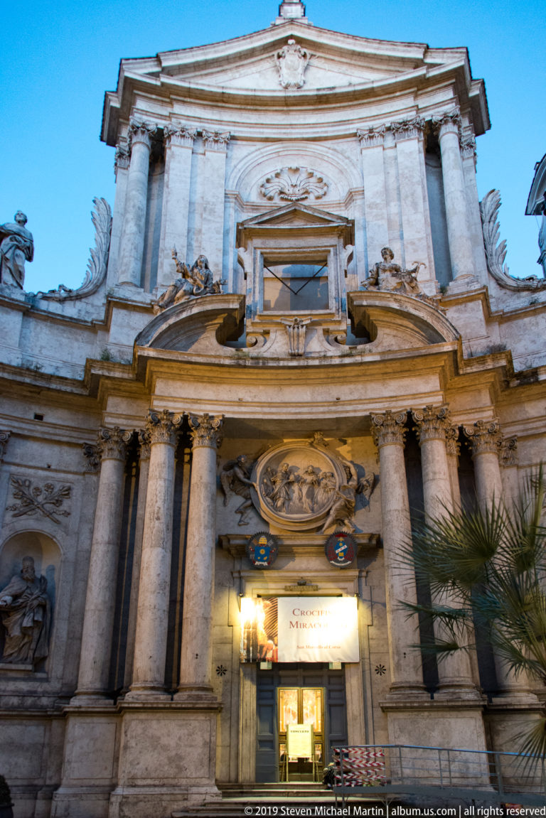 Upward view of an illuminated Baroque church façade with paired columns, pediments, sculptural details, and bright entrance.