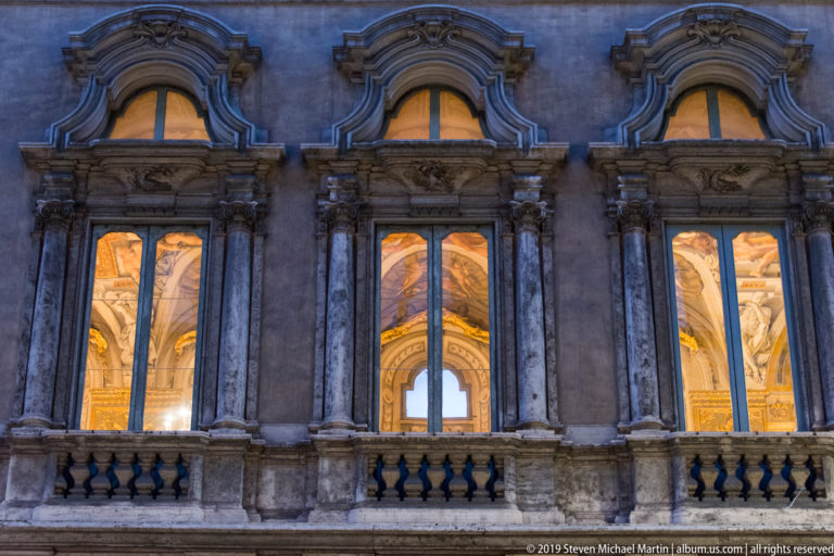Three tall illuminated arched windows in an ornate stone façade, with curved pediments, pilasters, balustrade, and painted interiors visible.