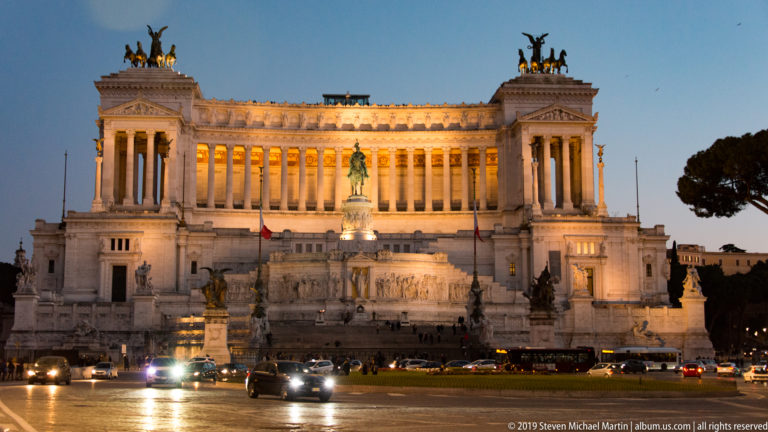 Wide illuminated Vittoriano at dusk with central statue and rooftop quadrigas above busy traffic in Piazza Venezia.
