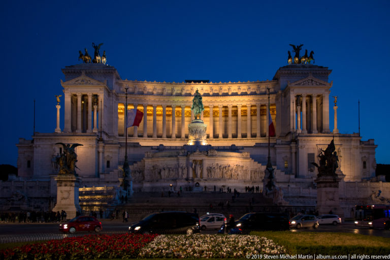 Illuminated Vittoriano at blue hour with central equestrian statue, rooftop quadrigas, flower beds, and cars crossing Piazza Venezia.