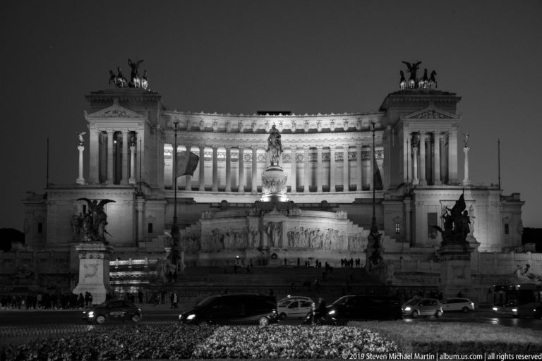 Black-and-white frontal night view of the illuminated Vittoriano, central equestrian statue, rooftop quadrigas, and traffic in front.