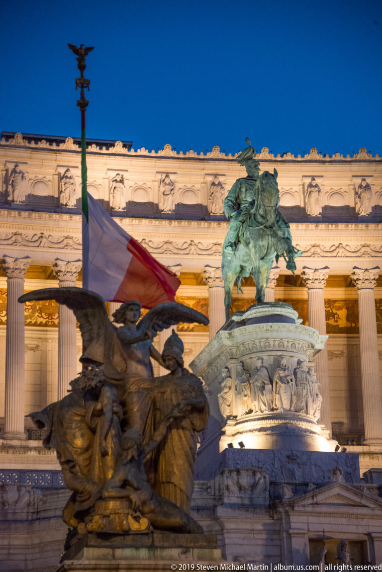 Bronze fountain sculpture before the Vittoriano, with Italian flag, equestrian statue, carved pedestal, and illuminated colonnade behind.