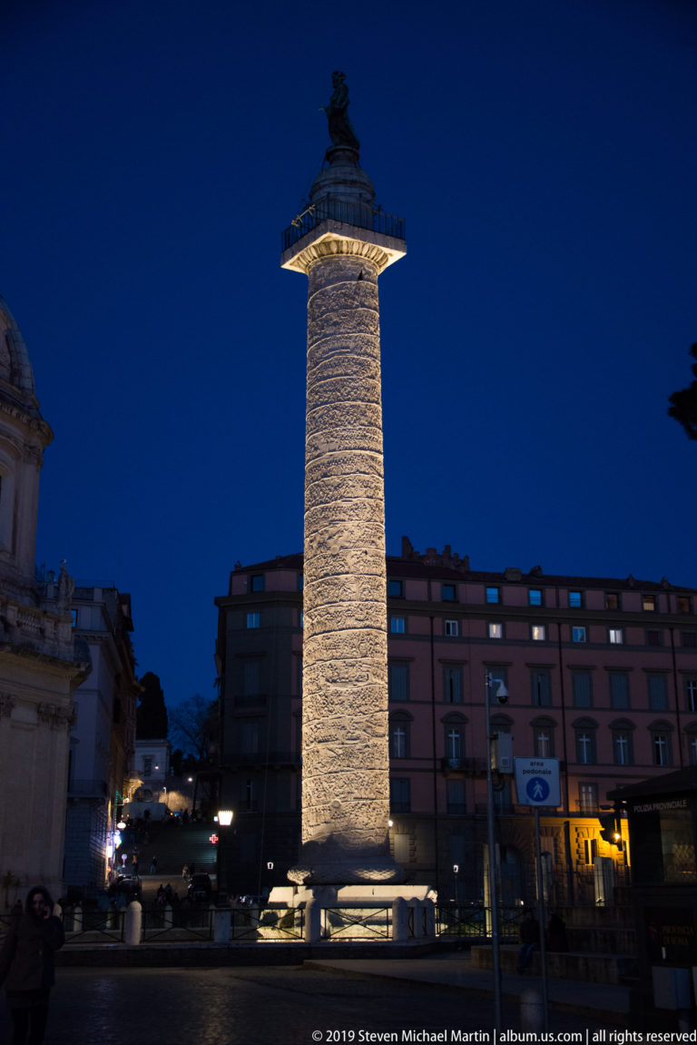 Full illuminated Trajan’s Column rising against a deep blue sky, with surrounding buildings and small figures at street level.
