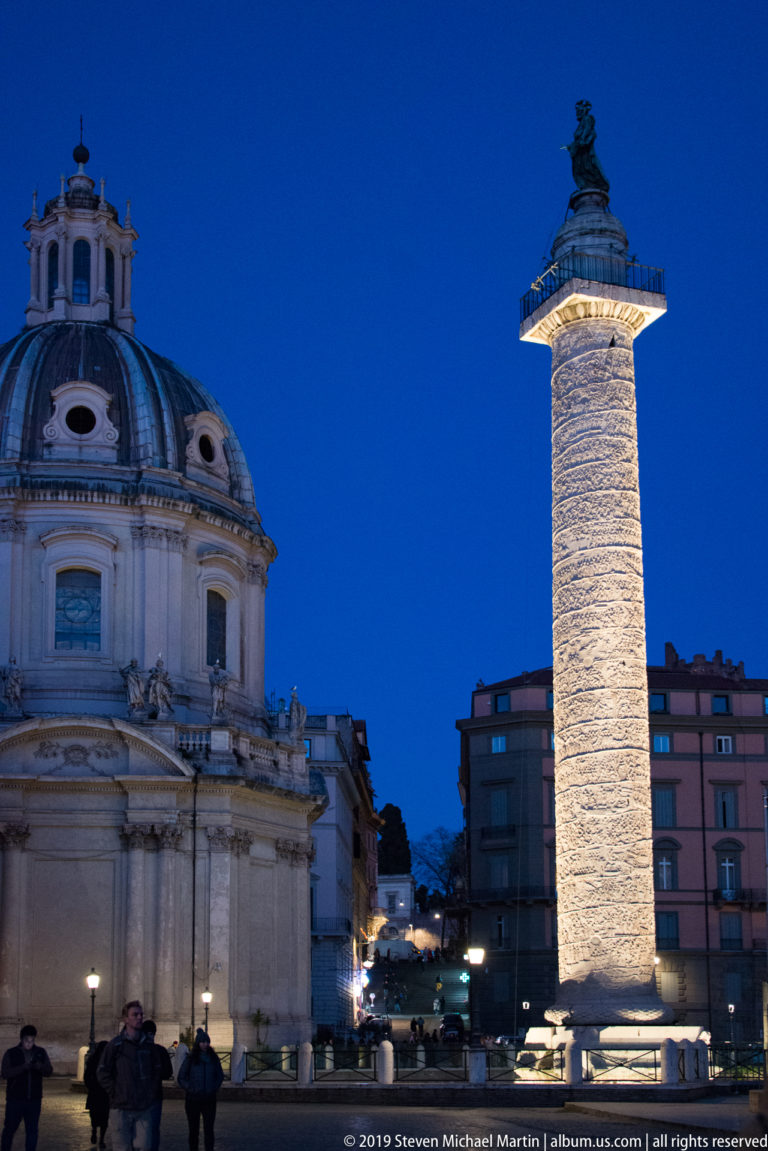 Trajan’s Column illuminated at dusk beside the domed Santa Maria di Loreto, with pedestrians and city buildings around the base.