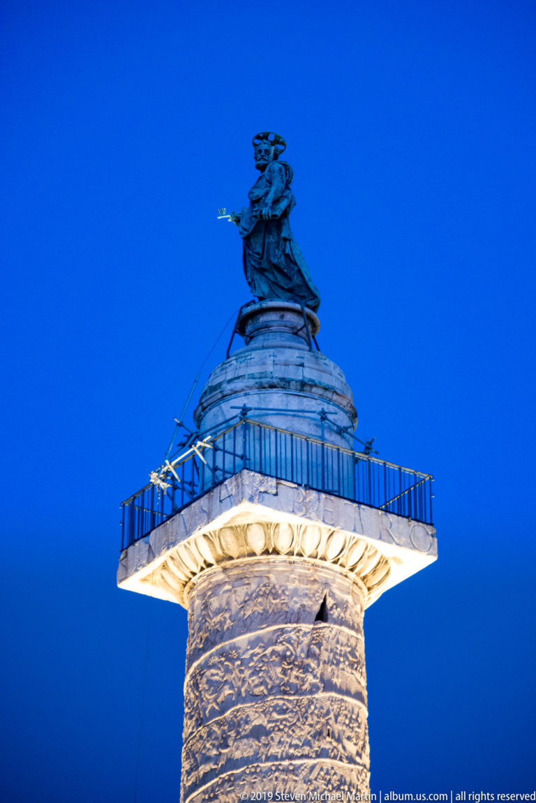 Illuminated upper shaft of Trajan’s Column with spiral reliefs, projecting capital, railing, and Saint Peter statue against blue evening sky.