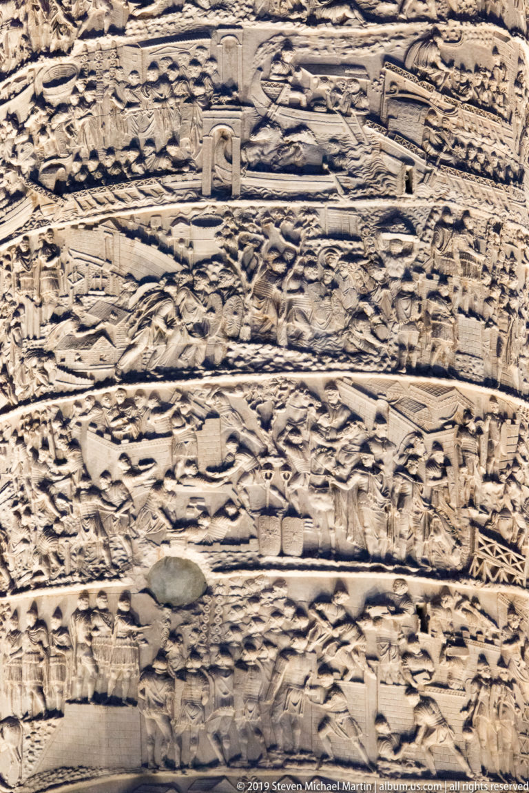 Tight illuminated detail of Trajan’s Column showing spiral relief bands with buildings, terrain patterns, walls, and small groups of figures.