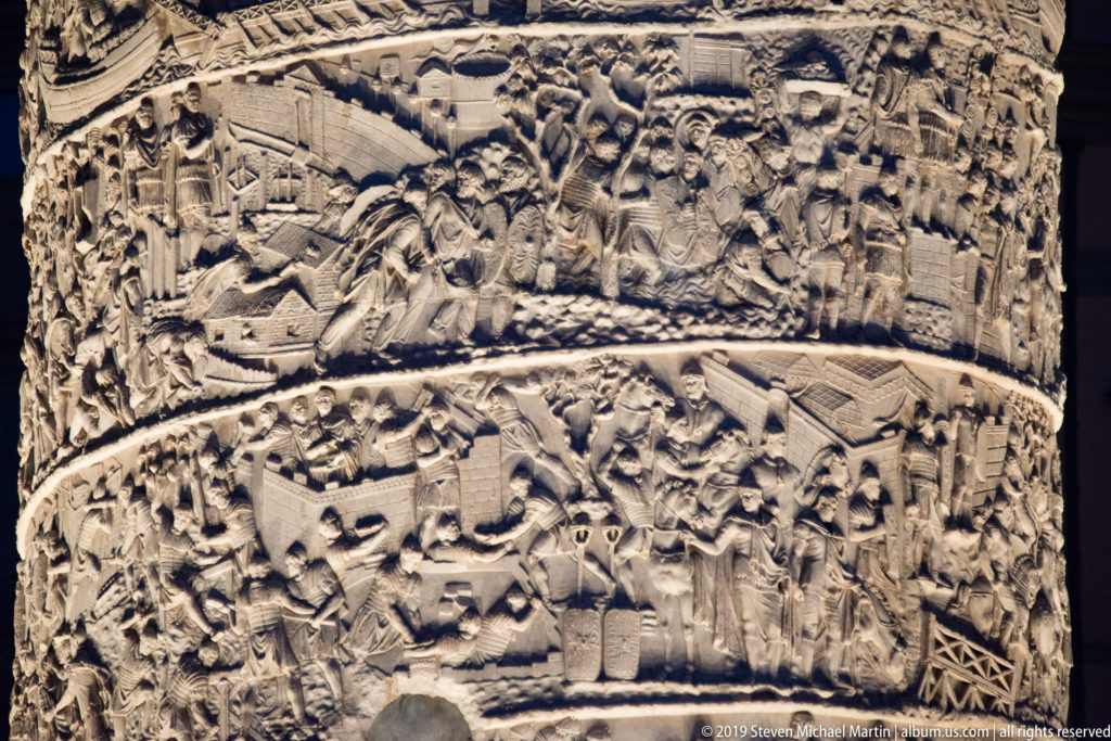 Night-lit detail of Trajan’s Column with crowded Roman figures, shields, fortifications, and construction or military activity across two spiral bands.