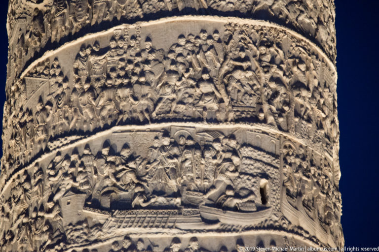 Close-up of two spiral bands on Trajan’s Column, with Roman soldiers, standards, buildings, and a carved boat picked out by side lighting.