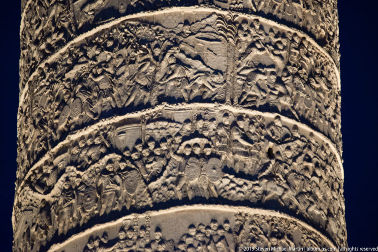 Close night detail of Trajan’s Column showing several spiraling bands of densely carved Roman soldiers, buildings, and landscape scenes.