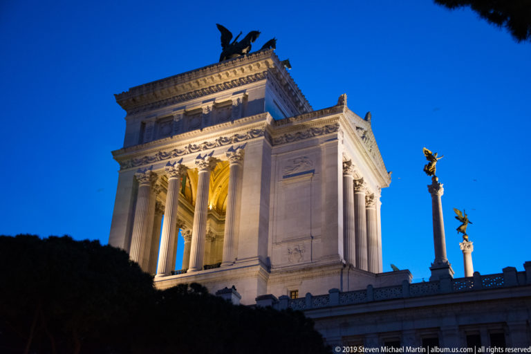 Low-angle night view of the illuminated Vittoriano, with its Corinthian portico, rooftop quadriga, and a gilded victory figure at right.