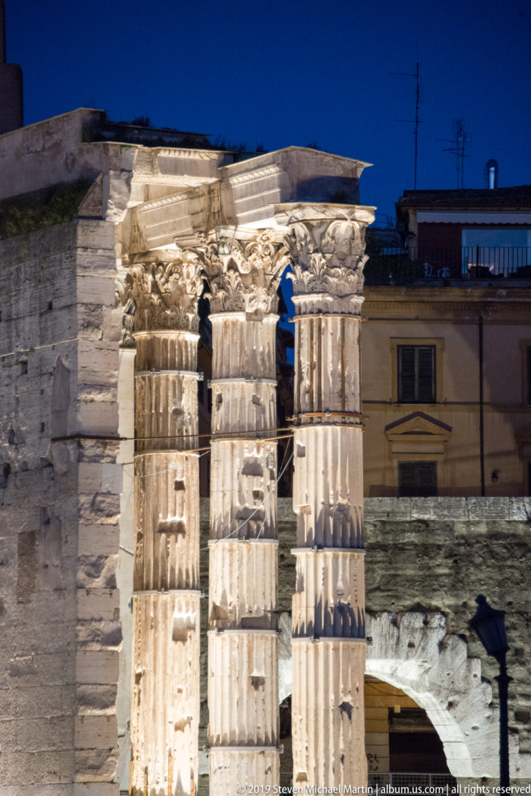 Floodlit fluted columns and carved entablature of the Colonnacce in the Forum of Nerva, with later buildings behind.