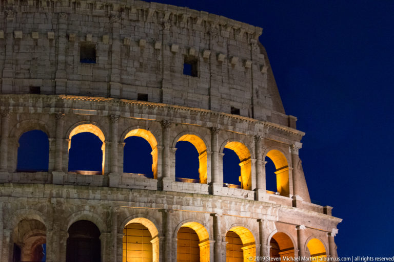 Frontal night view of the Colosseum with illuminated middle arcades, darker lower bays, and the massive surviving upper wall.