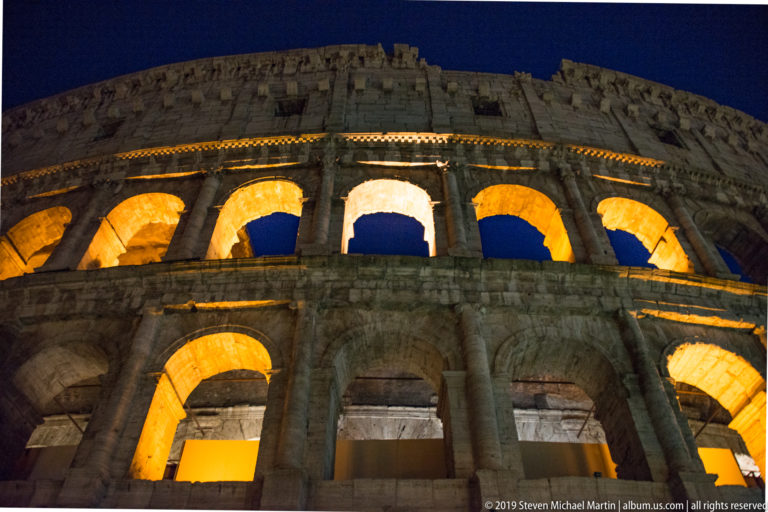 Tight upward night view of the Colosseum’s upper tiers, with amber-lit arches beneath the weathered surviving top wall.