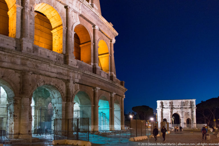 Colosseum arcades at left and the illuminated Arch of Constantine ahead, with pedestrians walking between the monuments at night.