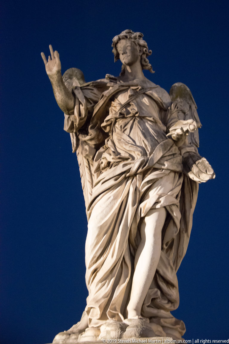 Low-angle view of a winged marble angel on Ponte Sant’Angelo carrying a large object amid deeply folded drapery.